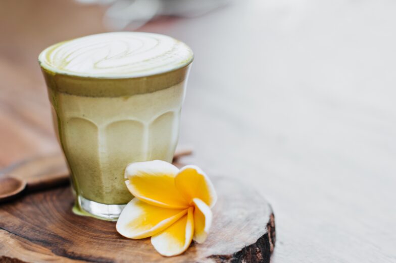 Glass of mocha matcha smoothie with coconut milk, served on a wooden board with a wooden spoon and a yellow flower beside it