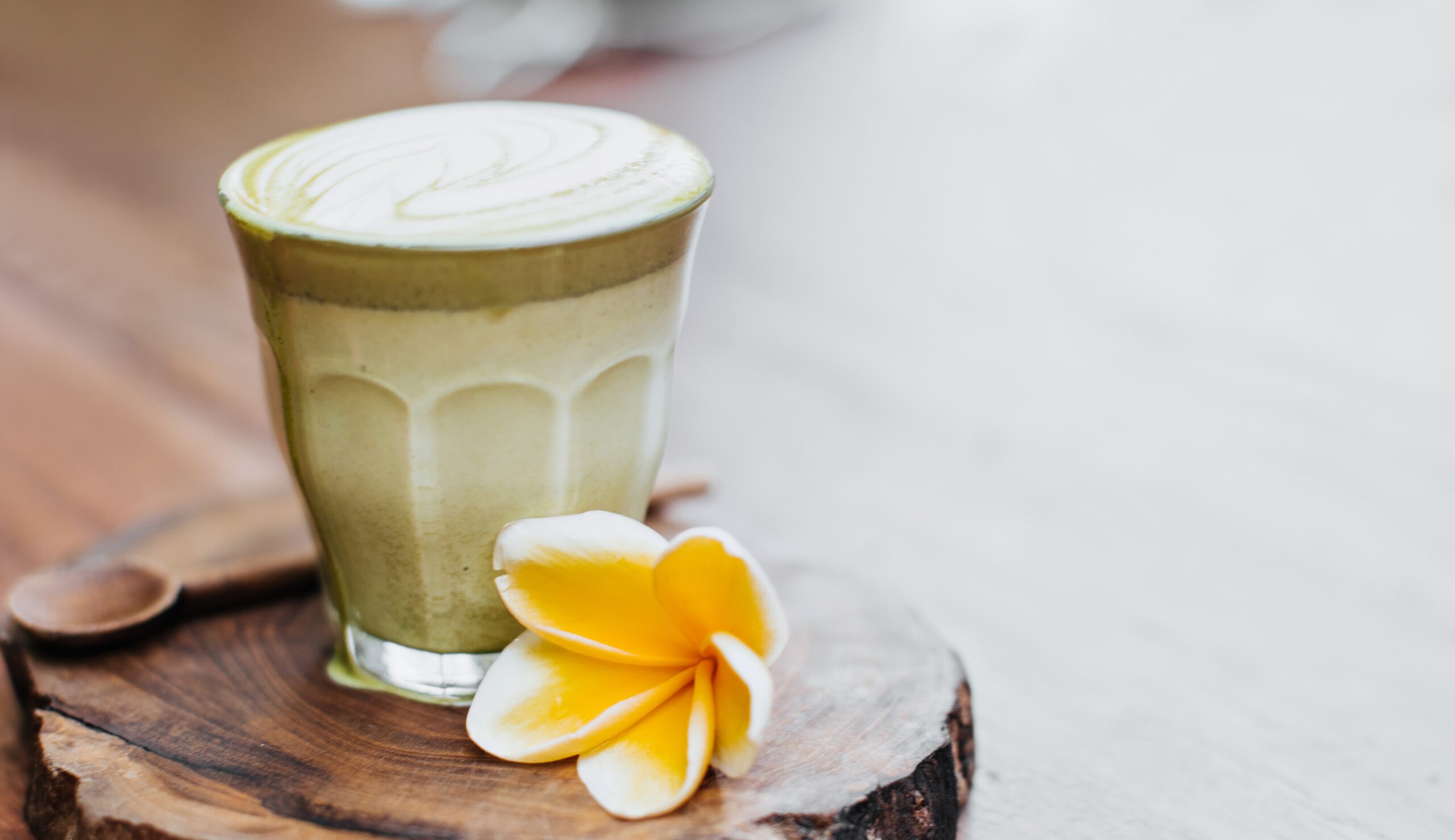 Glass of mocha matcha smoothie with coconut milk, served on a wooden board with a wooden spoon and a yellow flower beside it