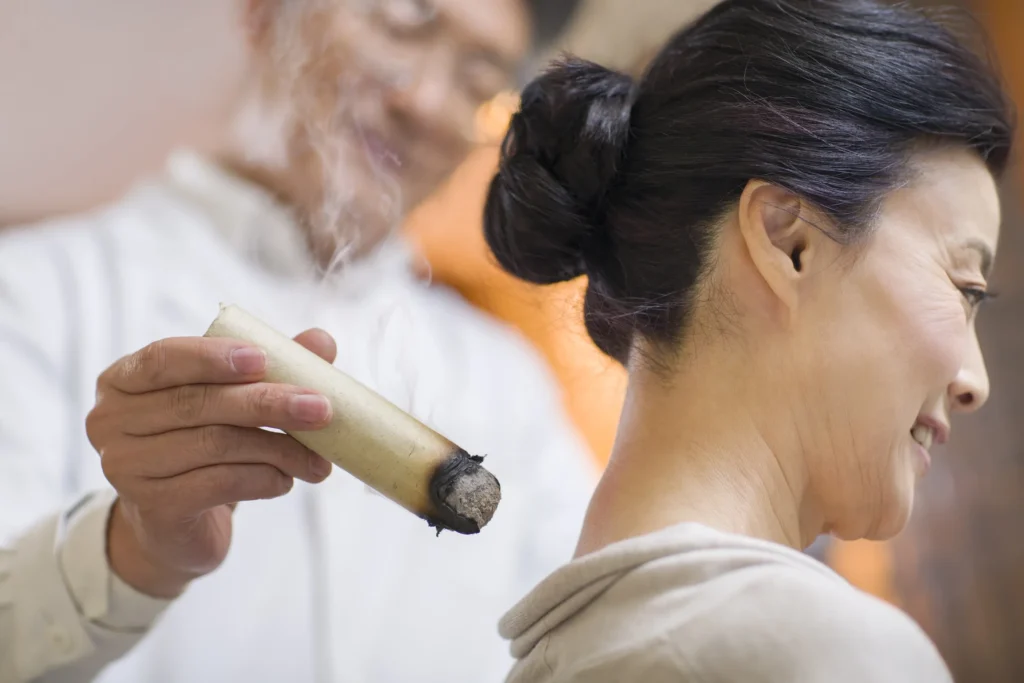Practitioner performing moxibustion by holding a lit moxa stick near the back of a woman’s neck during a traditional healing session.