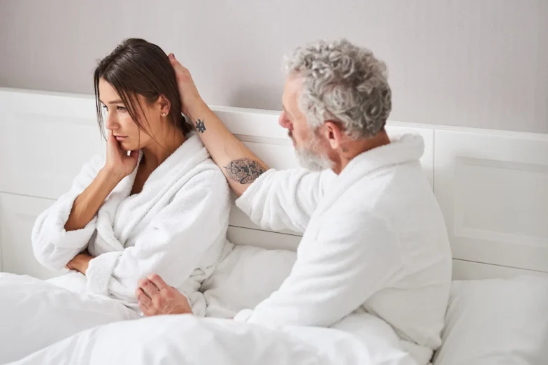 A woman sits on a bed in a white robe, looking away, while a man beside her gently touches her hair.