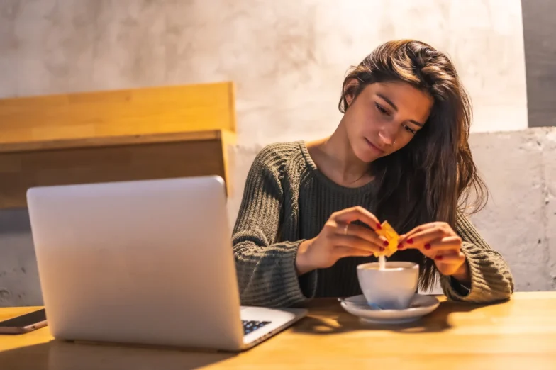 Woman at a table adding sweetener to a cup of coffee beside an open laptop