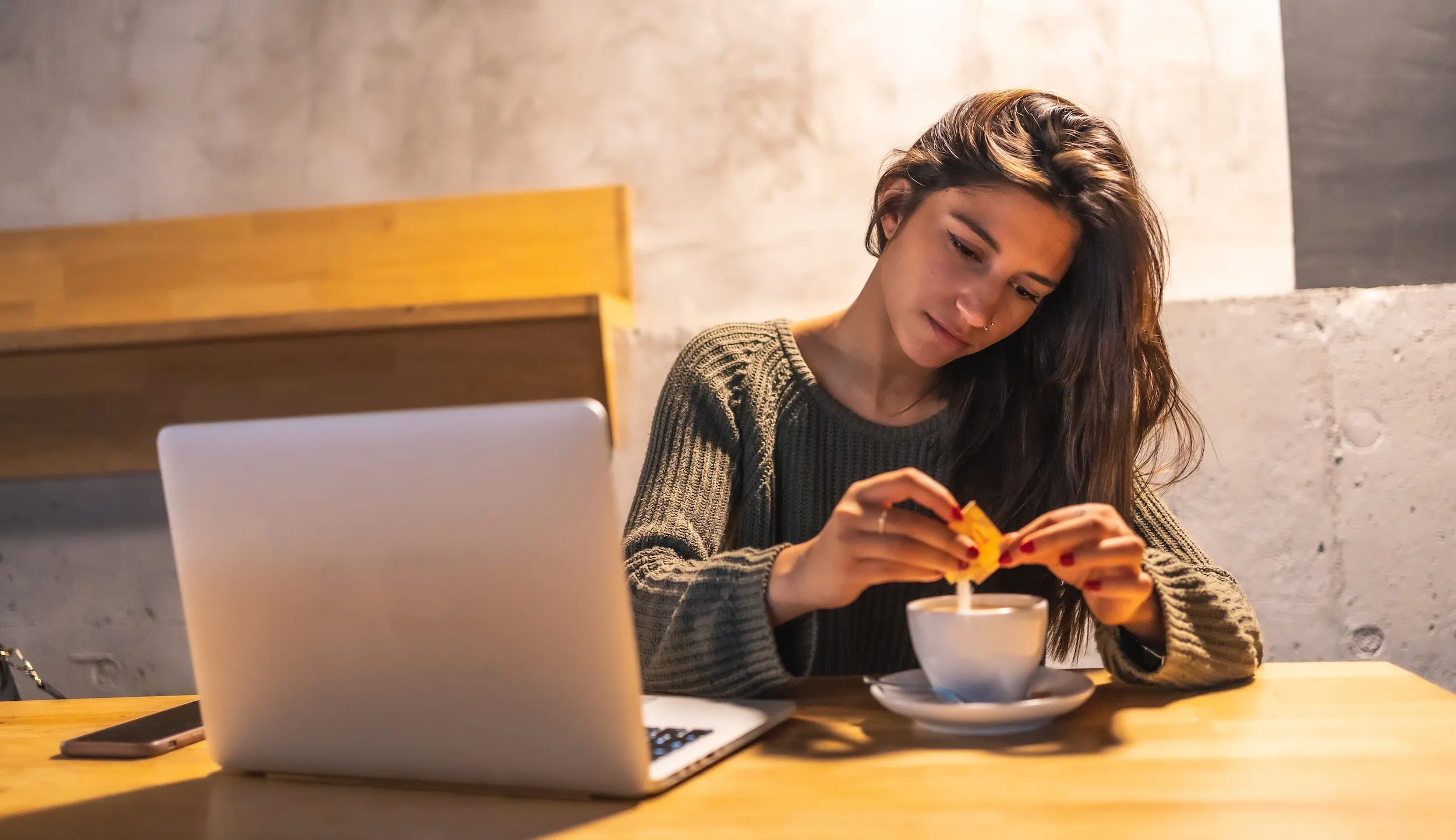 Woman at a table adding sweetener to a cup of coffee beside an open laptop