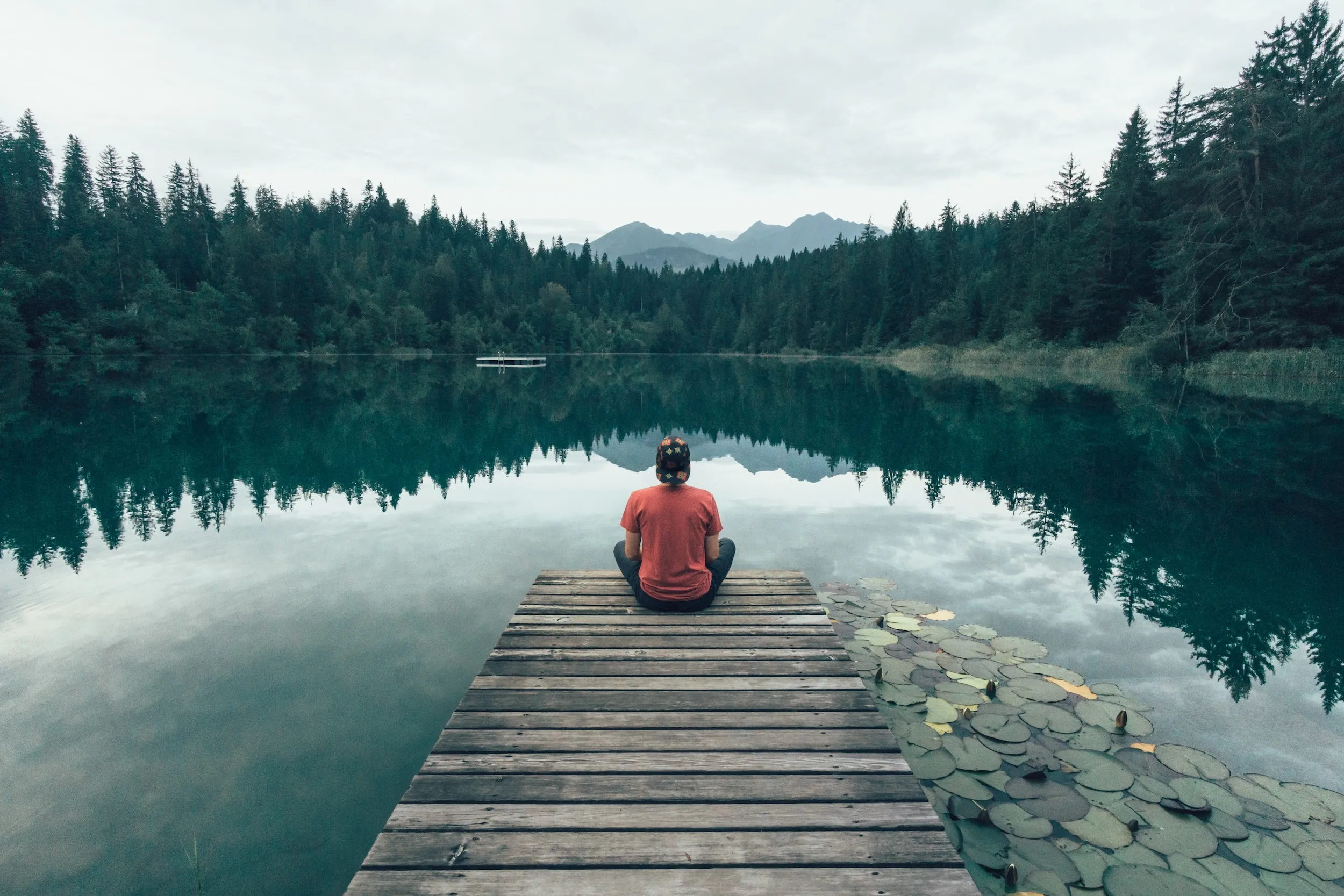 A person sitting quietly at the end of a wooden dock, facing a calm forest lake with mountains reflected in the water.