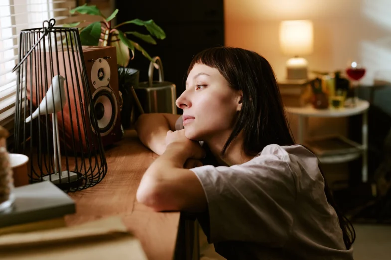 Woman resting her head and arms on a shelf indoors, gazing thoughtfully toward a window in a softly lit room.