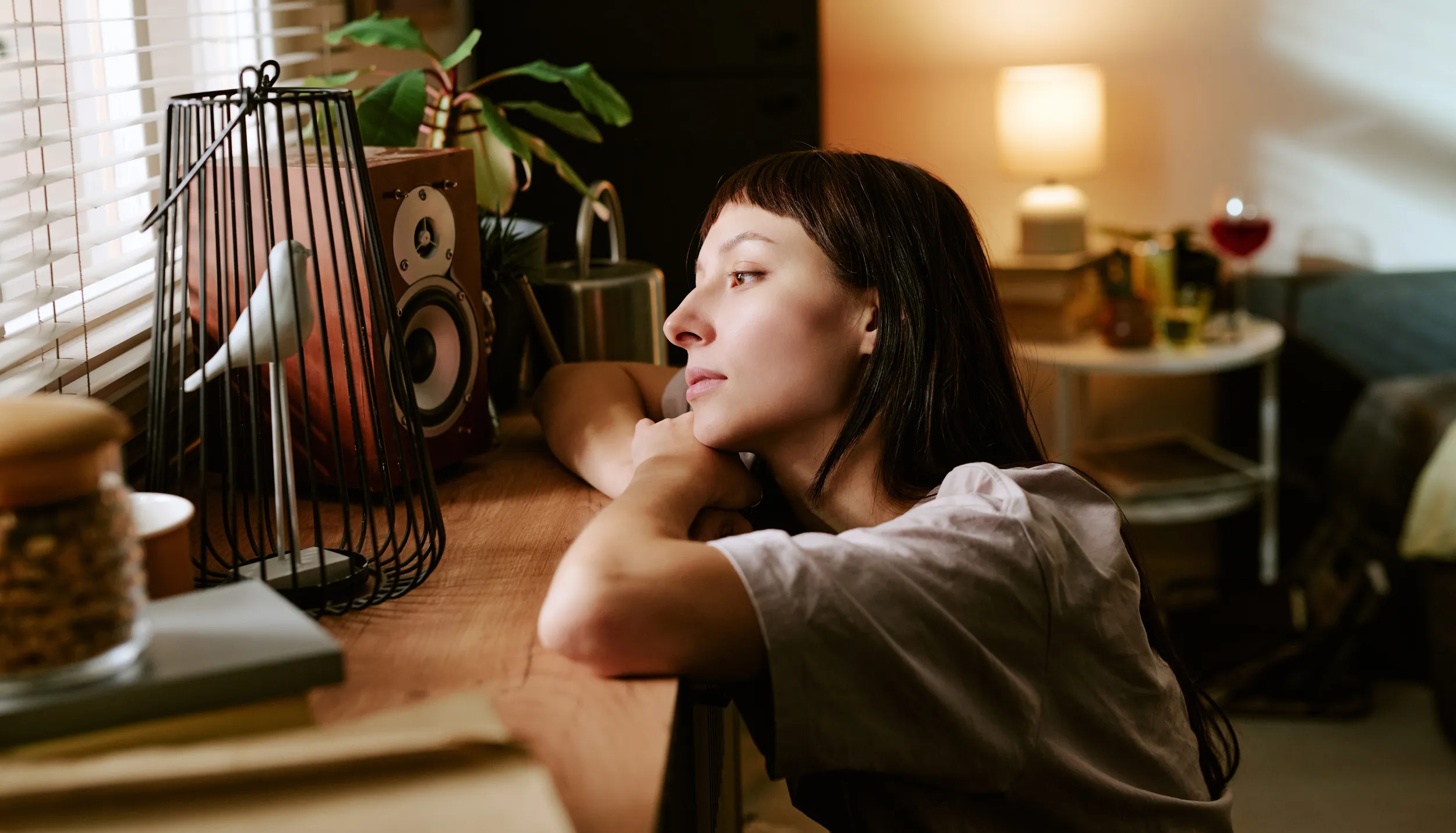 Woman resting her head and arms on a shelf indoors, gazing thoughtfully toward a window in a softly lit room.