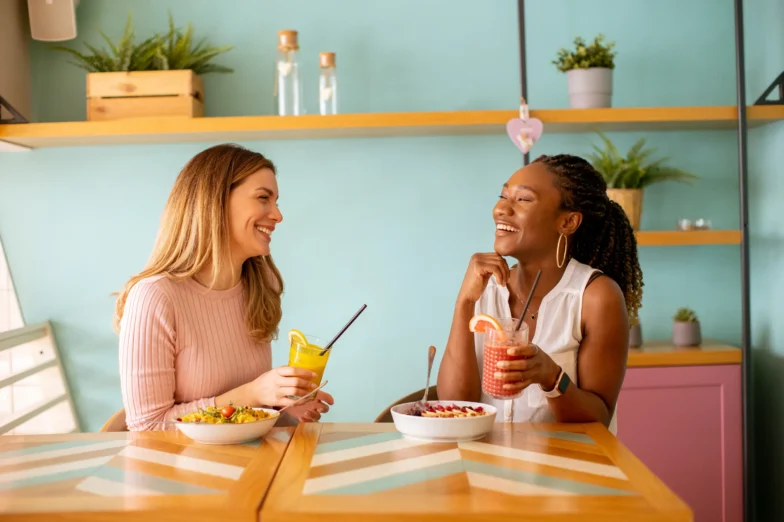 Two women sitting at a table, smiling and talking while sharing a healthy meal and drinks.
