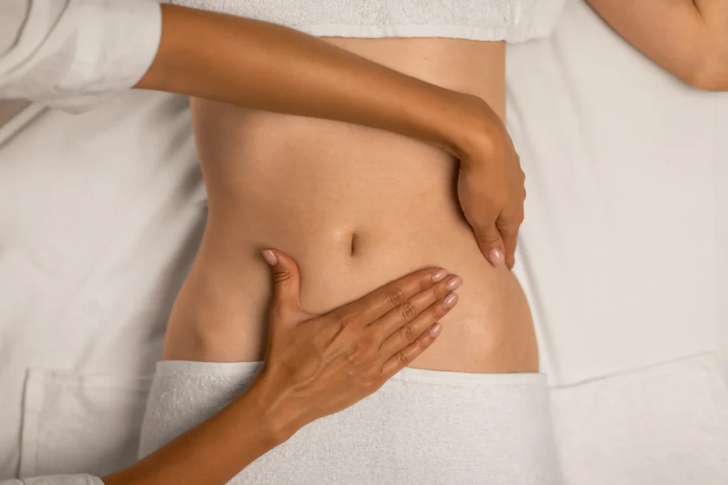 Close-up of a practitioner’s hands performing manual therapy on a woman’s lower abdomen while she lies on a treatment table.