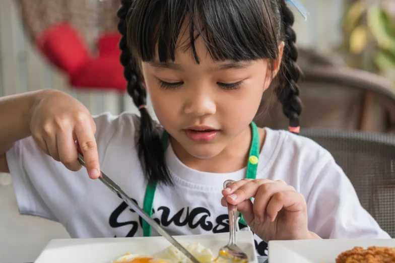 Young girl with braided hair eating a fried egg at a table, focusing carefully on her plate.