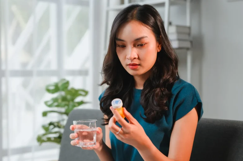 Woman sitting on a couch holding a prescription medication bottle and a glass of water.