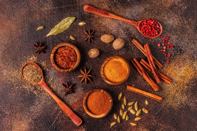 Assortment of culinary herbs and spices, including turmeric, cinnamon, cardamom, star anise, cumin, and pink peppercorns arranged on a rustic background.