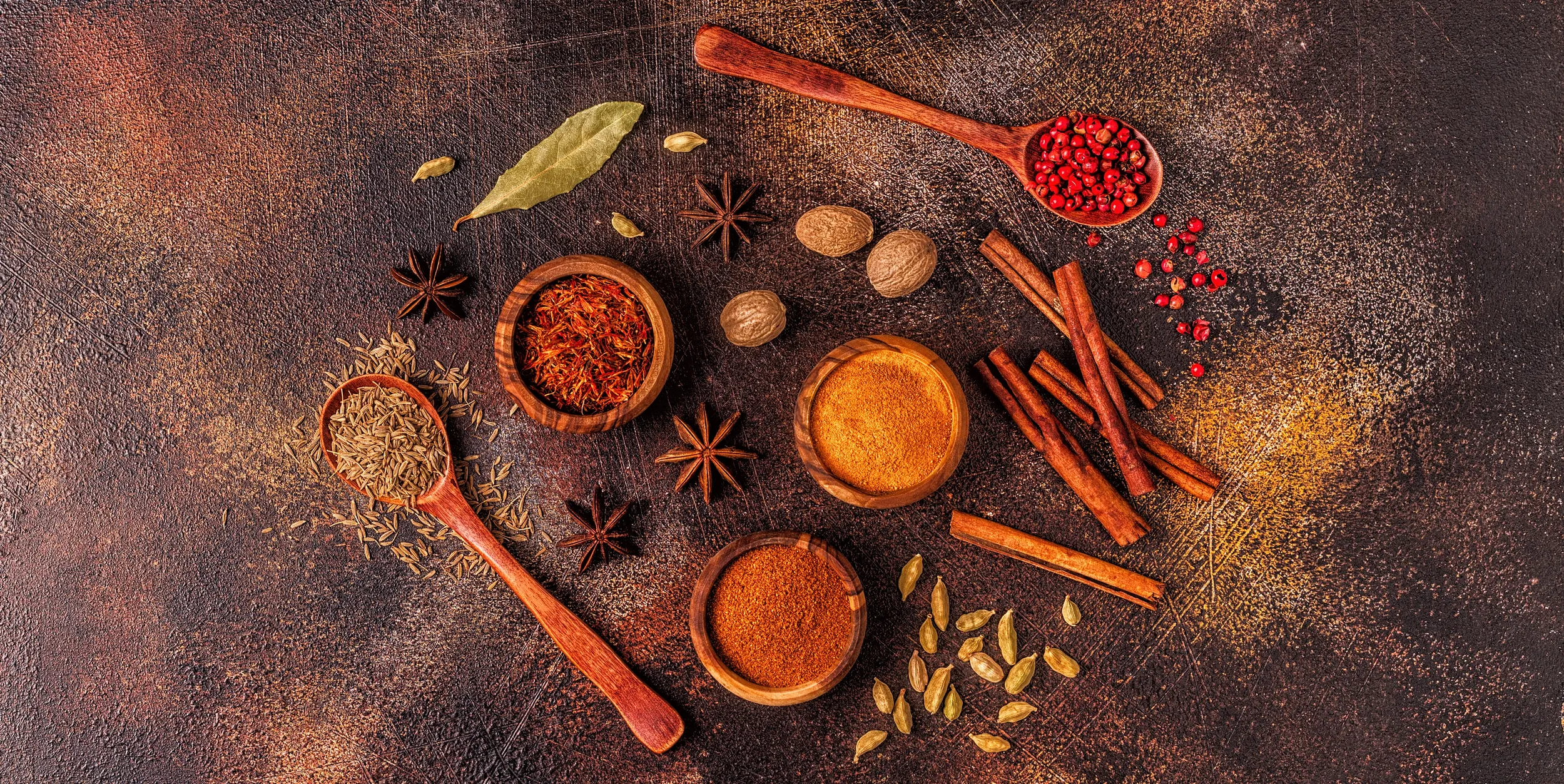 Assortment of culinary herbs and spices, including turmeric, cinnamon, cardamom, star anise, cumin, and pink peppercorns arranged on a rustic background.