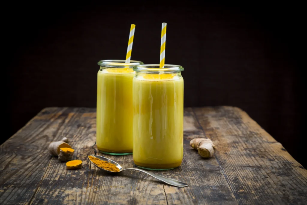 Golden turmeric smoothie in glass jars with straws, surrounded by fresh turmeric root and turmeric powder on a rustic wooden table.