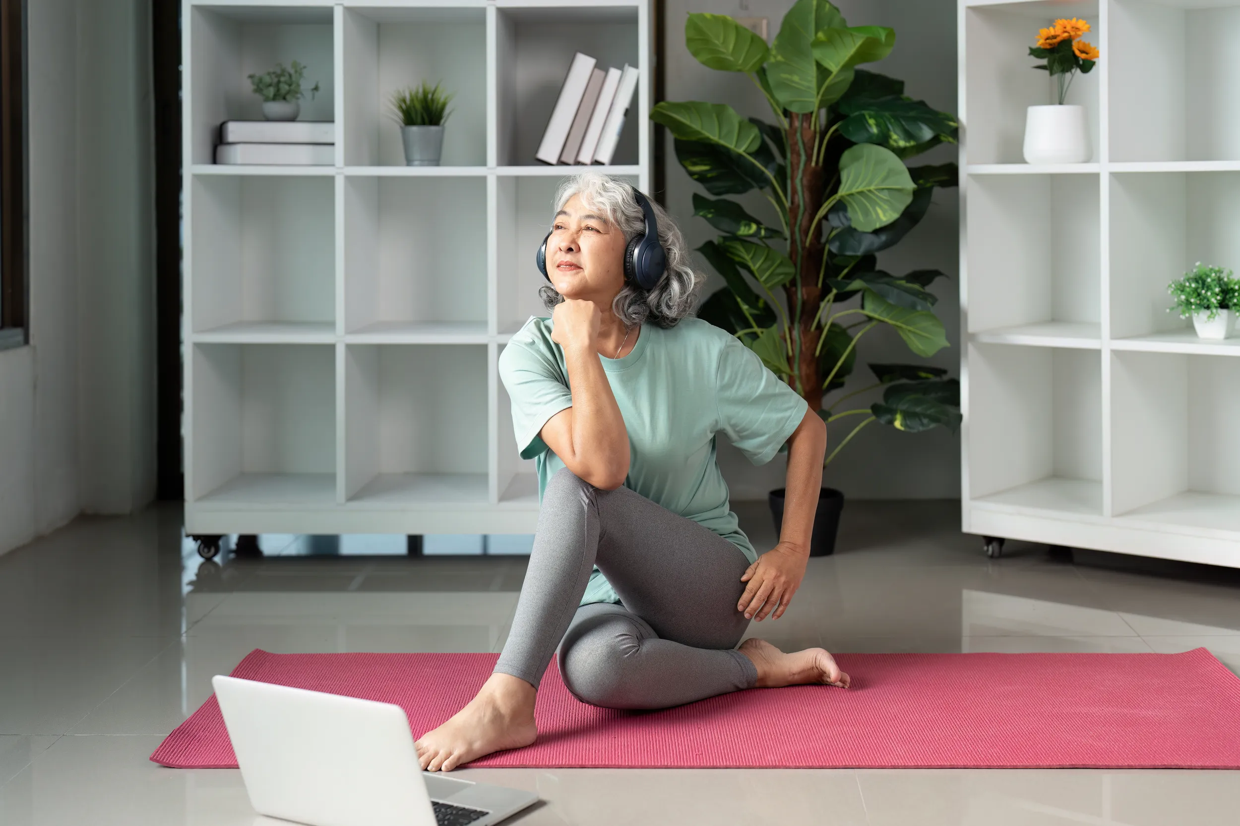 Older adult woman sitting on a yoga mat at home, wearing headphones and practicing a gentle seated stretch while following a session on a laptop.