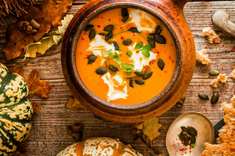 Rustic bowl of orange soup topped with pumpkin seeds, cream, and herbs, surrounded by autumn leaves and small pumpkins on a wooden table.