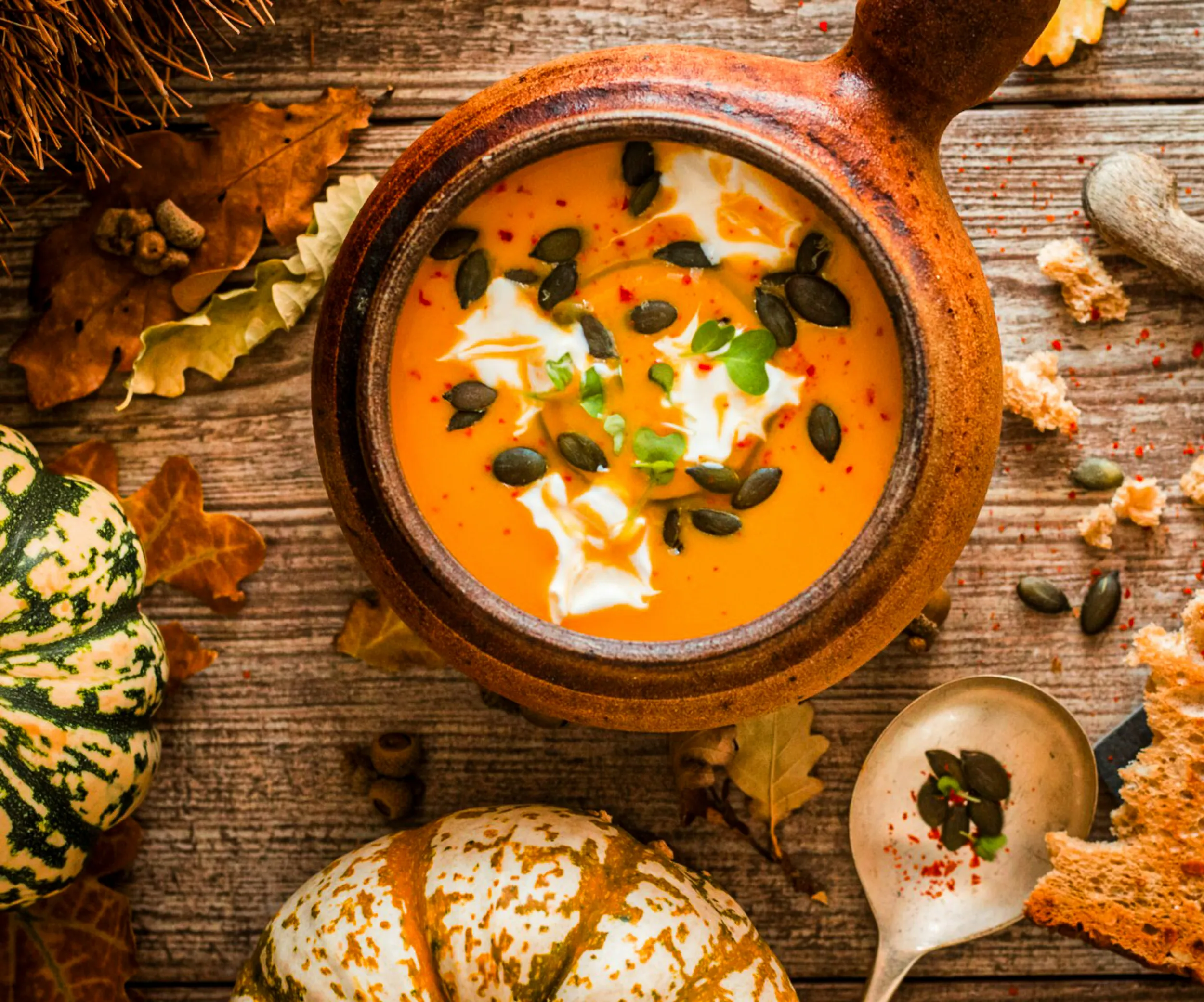 Rustic bowl of orange soup topped with pumpkin seeds, cream, and herbs, surrounded by autumn leaves and small pumpkins on a wooden table.