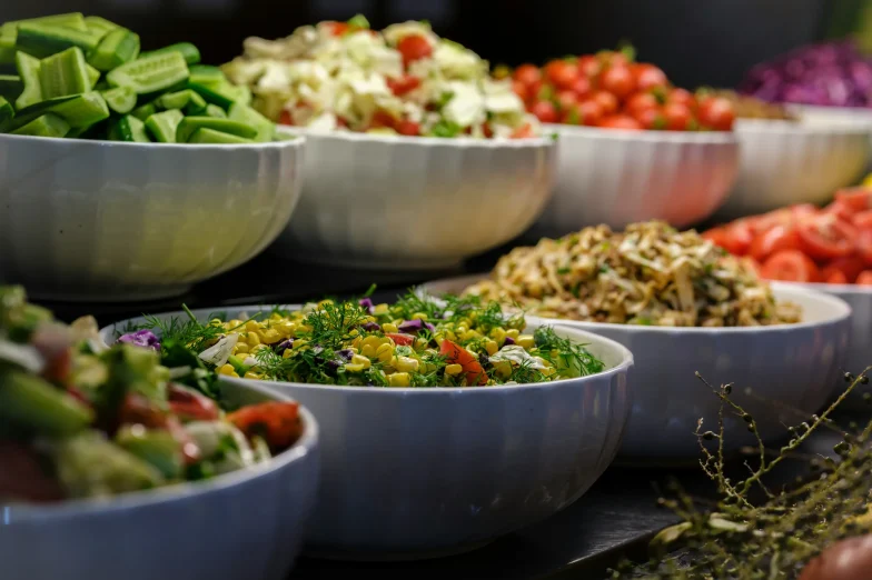 Assortment of colorful, nutritious salads and fresh vegetables in white bowls, featuring cucumbers, tomatoes, corn, leafy greens, and herbs.